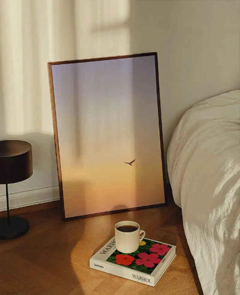 Minimalistyczny druk artystyczny w drewnianej ramie przedstawiający samotnego ptaka w locie na tle delikatnego gradientu nieba przechodzącego od bladego lawendowego do ciepłego pomarańczowego.