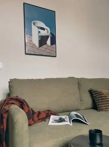 Oliwkowa zielona kanapa obita tkaniną z wzorzystym pledem zarzuconym na podłokietnik i paskowym poduszką spoczywającą na siedzisku